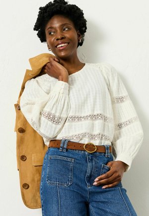 Smiling woman with short curly hair wearing a white lace blouse, blue jeans with brown belt, and holding a tan jacket over her shoulder.