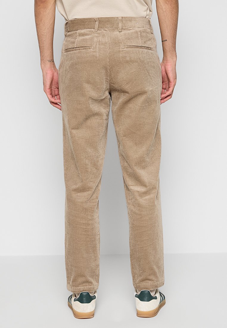Braune Cordhose mit geradem Schnitt, strukturierter Oberfläche, zwei Gesäßtaschen und einem Bund. Getragen mit grünen und weißen Turnschuhen.