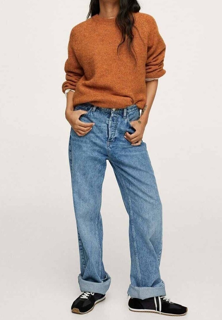 Femme portant un pull orange rouille, un jean bleu ample avec revers, et des baskets noires, debout les mains dans les poches sur un fond uni.
