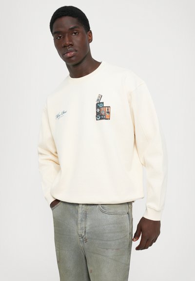 Homem com um sweatshirt creme com um gráfico de alto-falante e jeans claros desbotados, de pé contra um fundo claro simples, com as mãos nos bolsos.