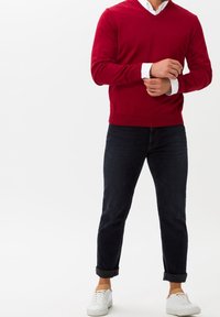 Pull rouge à col en V en tissu doux, porté sur une chemise blanche. Associé à un jean en denim foncé et des baskets blanches, avec des poignets retroussés.