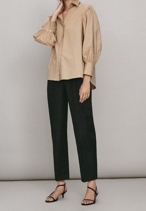Femme portant un chemisier beige à manches longues bouffantes, un pantalon noir longueur cheville et des sandales noires à brides et talons bas, debout à l'intérieur.