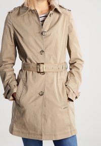 Beige trenchcoat met een riem in de taille, schouderepauletten en knoopsluiting. Twee zijzakken en een gladde, gestructureerde stof.