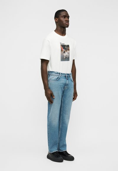 Λευκό βαμβακερό μπλουζάκι με φωτογραφικό τύπωμα, συνδυασμένο με ανοιχτόχρωμο τζιν παντελόνι. Το μοντέλο φοράει μαύρα παπούτσια με κορδόνια. Απλός σχεδιασμός, άνετη εφαρμογή.
