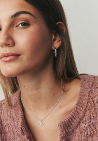 Collana d'argento con cuori intrecciati, catena delicata, abbinata a un orecchino a cerchio d'argento. Il modello indossa un maglione rosa lavorato a maglia con trama.