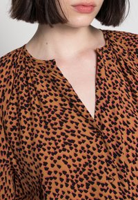 Blusa marrone con stampa a pois neri e rosa, caratterizzata da una scollatura a V e dettagli arricciati sulle spalle. Tessuto morbido.