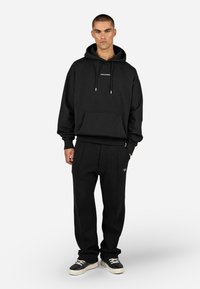 Μαύρη φούτερ με κουκούλα και μπροστινή τσέπη, συνδυασμένη με υφασμάτινους μαύρους παντελόνες με λογότυπο. Το σύνολο ολοκληρώνεται με μαύρα αθλητικά παπούτσια.