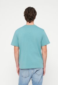 Turkio spalvos medvilninė marškinėliai su apvalia iškirpte ir trumpomis rankovėmis, pasižymintys paprasta, lygiu tekstūra, dėvimi su šviesiai mėlynomis džinsomis.