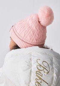 Lyserød strikket hue med et tekstureret mønster, pyntet med små perler og en fluffy pompon på toppen. Bruges sammen med en hvid dunjakke.