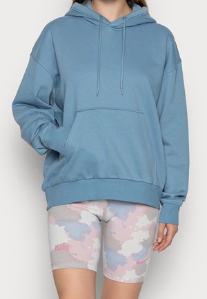 Osoba ubrana w niebieską, oversize bluzę z kapturem z przednią kieszenią oraz pastelowe, różowe, niebieskie i beżowe krótkie spodenki rowerowe Nike z wzorem.