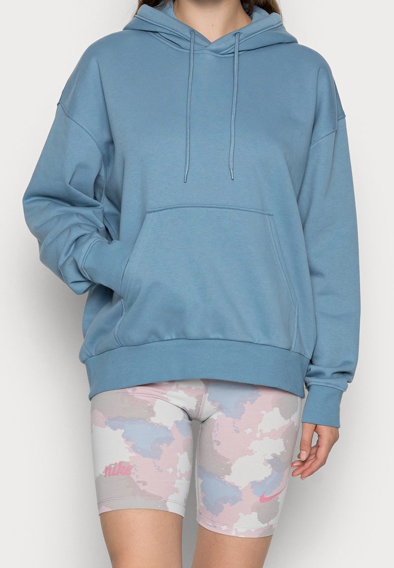 Personne portant un sweat à capuche bleu oversize avec poche avant et un short de cycliste Nike à motifs pastel rose, bleu et beige.