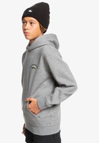 Sudadera con capucha gris hecha de una mezcla de algodón, con un bolsillo frontal, puños acanalados y el logo de Quiksilver en bordado verde y amarillo. Lleva un gorro negro.