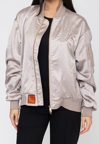Femme portant une veste bomber en satin beige clair avec une fermeture éclair dorée, un « B » brodé sur la poitrine, une poche sur la manche et une étiquette orange près de l’ourlet.