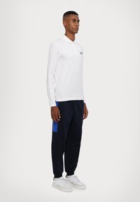 Hvit langermet polo med svart logo, kombinert med marineblå joggebukser med blå sidelapp og hvite joggesko.