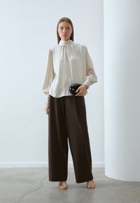 Kremfarget silkebluse med puff ermer, høy krage og elastiske mansjetter; brune vidde bukser; holder en sort oval clutch; gullfargede sandaler med stropper.