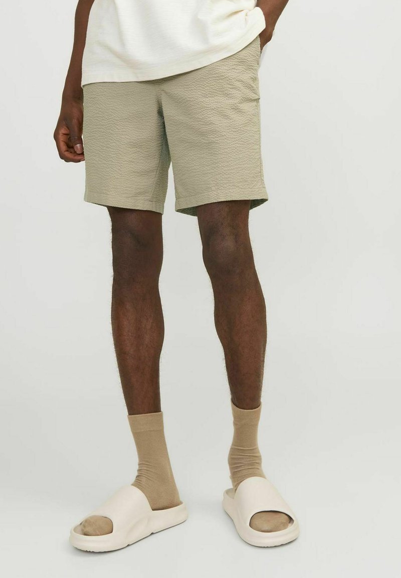 Haki šorti ar teksturētu audumu, virs ceļgaliem, kopā ar bēšām zeķēm un gaismas krāsas čībām. Vienkāršs dizains, bez rakstiem.