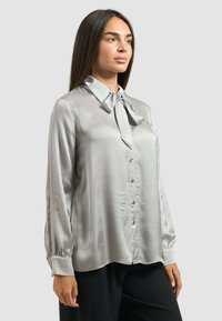 Blusa leggera di seta grigia chiara con colletto a punta e dettaglio del fiocco, caratterizzata da bottoni sulla parte frontale e maniche arricciate.