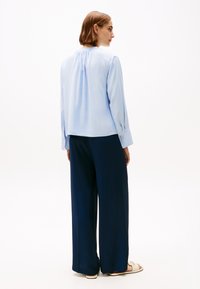 Blusa azzurra a maniche lunghe con retro plissettato, abbinata a pantaloni a gamba larga blu navy. I pantaloni presentano una apertura laterale e la blusa ha polsini con bottoni.