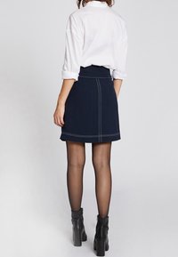 Granatowa spódnica dżinsowa z białymi szwami, w kształcie litery A, noszona z białą koszulą z kołnierzykiem i czarnymi botkami za kostkę, widok z tyłu.