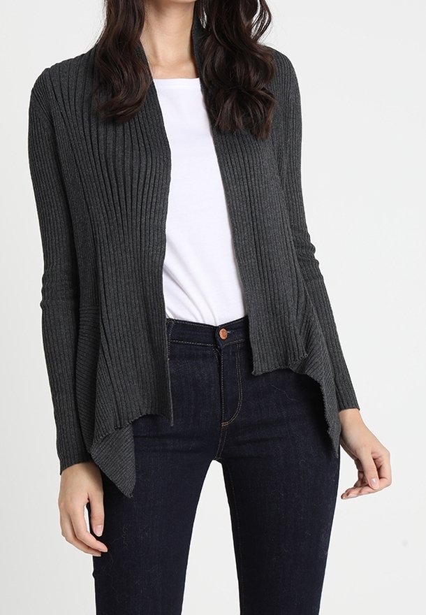 Femme portant un cardigan gris foncé à côtes et ouvert sur le devant, par-dessus une chemise blanche et un jean bleu foncé, visible du cou aux hanches sur un fond uni.