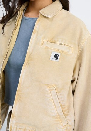 Beige Carhartt jakke med lynlås, brystlomme og logo-patch båret over en blå ribbet top.