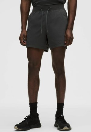 Persona atletica che indossa pantaloncini neri con coulisse, calzini neri e scarpe da corsa nere, in piedi su uno sfondo chiaro e uniforme.