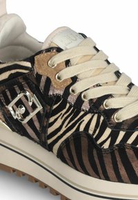 Zapatilla con estampado de cebra en negro, marrón y crema, que presenta herrajes metálicos, cordones color crema y una suela de goma con un patrón a rayas.