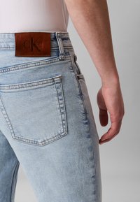 Gaiši zili džinsa džinsi ar brūnas krāsas Calvin Klein ādas logo uzlikumu pie jostasvietas, ar divām muguras kabatām un taisnu kāju dizainu.