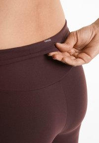 Burgundy high-waisted leggings with a smooth texture and a logo on the waistband; hand gripping the fabric near the waistband.