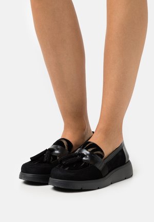 Chaussures noires à enfiler, combinant cuir brillant et daim doux, ornées de glands décoratifs et d'une semelle noire épaisse.