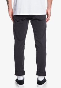 Pantalon en coton gris foncé avec une coupe droite, ourlets retroussés et poches arrière. Porté avec un t-shirt blanc et des baskets noires.