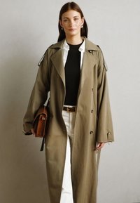 Cappotto trench khaki con ampi revers, grandi bottoni e spalline, abbinato a una maglietta nera e pantaloni bianchi. Bicchiere marrone come accessorio.