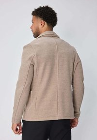Homme aux cheveux bouclés courts portant une veste beige vu de dos sur un fond gris clair uni.