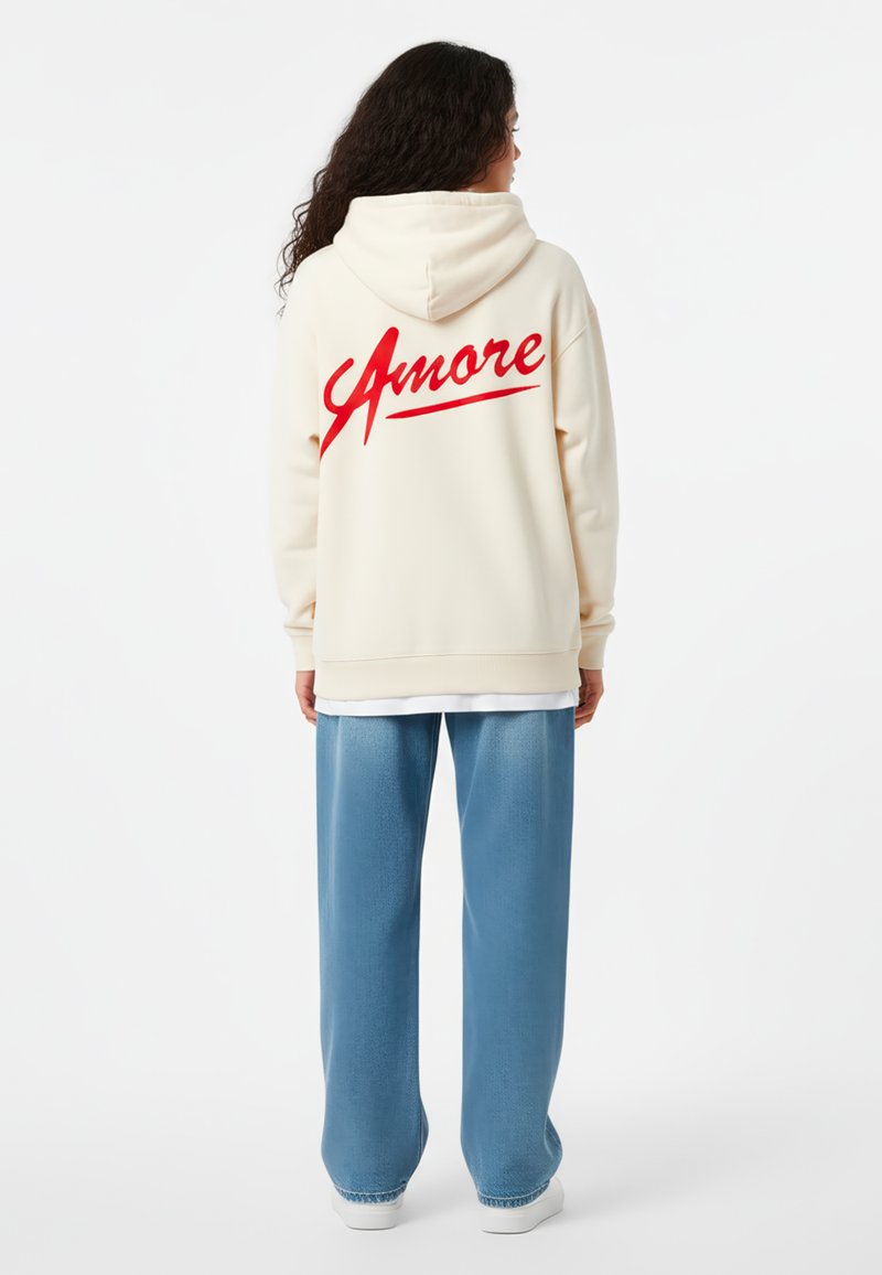 Kremast hoodie s crvenim natpisom "Amore" na leđima, u kombinaciji s svijetloplavim hlačama širokog kroja. Glatka tkanina, opušteni kroj, s kapuljačom.
