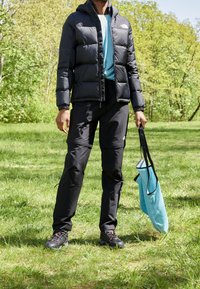 Svart pufferjacka, blå skjorta, svarta byxor, svarta skor, håller en ljusblå tygväska, står på grön gräs med träd i bakgrunden.