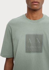 Světle zelené tričko s krátkým rukávem vyrobené z jemné bavlny, na přední straně s potiskem čtvercového loga v kontrastním tmavém vzoru.