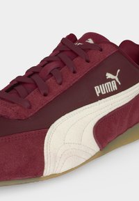 Baskets Puma marron foncé avec bande latérale blanche, dessus en suède et tissu, lacets marron foncé et semelle en gomme.