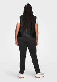 Černá kožená vesta, bílá košile s krátkým rukávem, tmavé džíny a světlé tenisky. Dlouhé copy splývají vzadu. Neutrální pozadí.