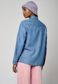 Camisa de denim en azul claro, con puños abotonados, dobladillo ligeramente más largo por detrás, combinada con pantalones rosas. Estilizada con un suave gorro morado de canalé.