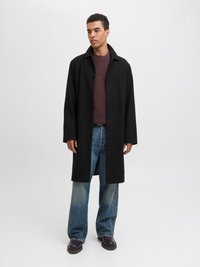 Άνδρας φορώντας μαύρο μακρύ παλτό, μπορντό πλεκτό πουλόβερ, μπλε τζιν και καφέ παπούτσια στέκεται μπροστά σε λευκό φόντο.