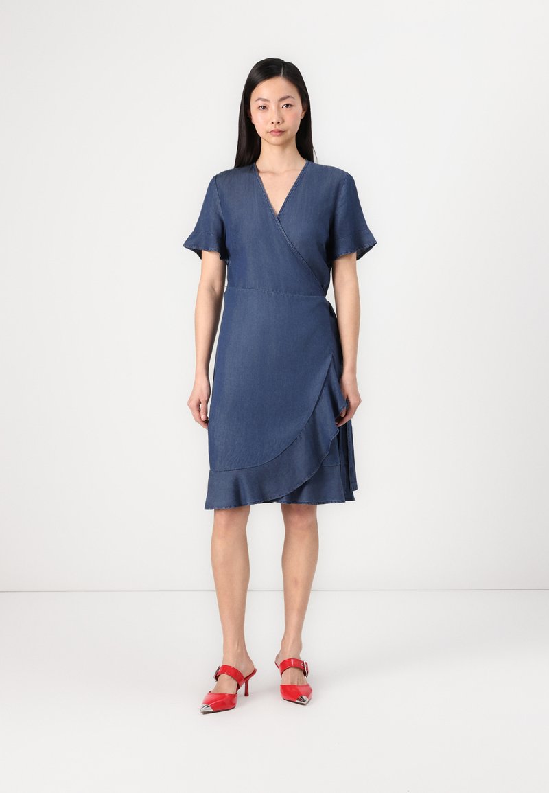 Robe chasuble en denim avec des manches courtes et évasées, décolleté en V et ourlet superposé. Femme portant des sandales à talons rouges à bouts pointus.