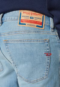 Jeans de denim azul claro con una etiqueta de cuero marrón que presenta gráficos en rojo y azul. Dos bolsillos traseros con detalles de costura sutil.