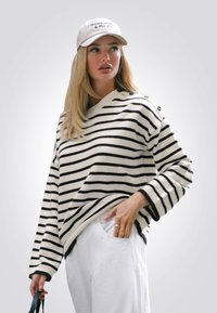 Maglione a righe in crema e nero, design oversize con dettagli a bottone sulle spalle, abbinato a pantaloni bianchi e un cappellino leggero.