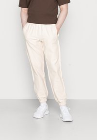 Pantalones jogger de color melocotón claro con cintura elástica y puños, que presentan una textura suave y un logotipo discreto en la pierna izquierda. Combinados con zapatillas blancas.