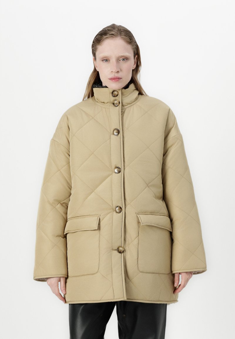 Beige prošívaná bunda s vysokým límcem, velkými předními kapsami a zapínáním na knoflíky. Má volný střih a hladkou texturu.