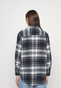 Plaid shirt featuring dark green, navy, white, and light pink patterns. Soft fabric, long sleeves, button cuffs, and curved hem. Back view.