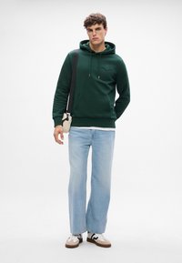 Felpa verde con cappuccio e tasca frontale, abbinata a jeans a gamba larga azzurri e sneakers bianche con dettagli neri e marroni.