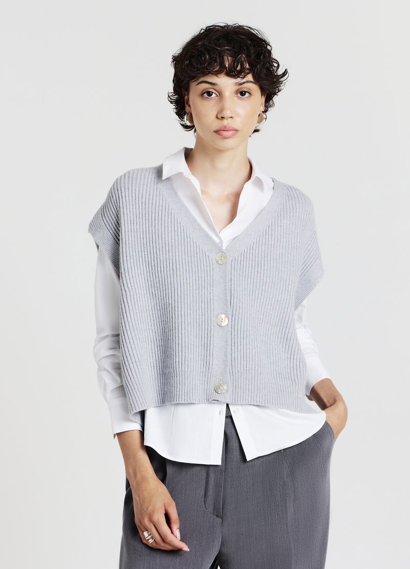 Débardeur court à côtes grises avec un col en V, comportant trois boutons. Porté sur une chemise blanche, mettant en avant une texture douce et une coupe décontractée.