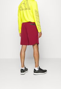 Pantalones cortos deportivos rojos con una textura suave, que cuentan con un bolsillo trasero con cremallera. Combinados con una camiseta de manga larga amarilla neón y zapatillas negras.