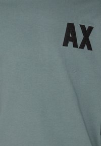 Bomull T-shirt i dämpad teal med en mjuk textur, med en svart broderad "AX" logotyp på vänster sida.